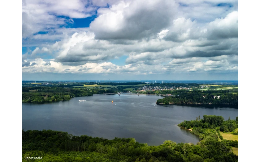 Luftaufnahme des Möhnesees mit Segelbooten und umliegender Landschaft unter Wolkenhimmel.