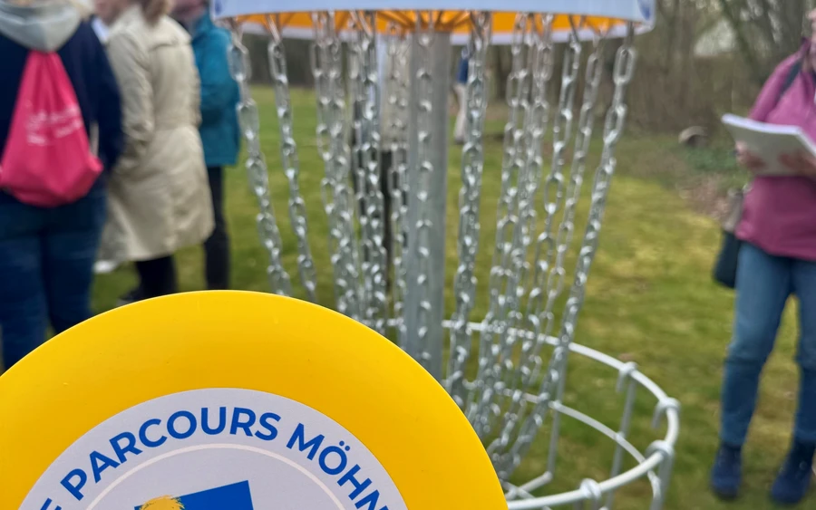 Discgolf-Scheibe mit Logo des Discgolf-Parcours Möhnesee vor einem Korb mit Schild „Staumauer"