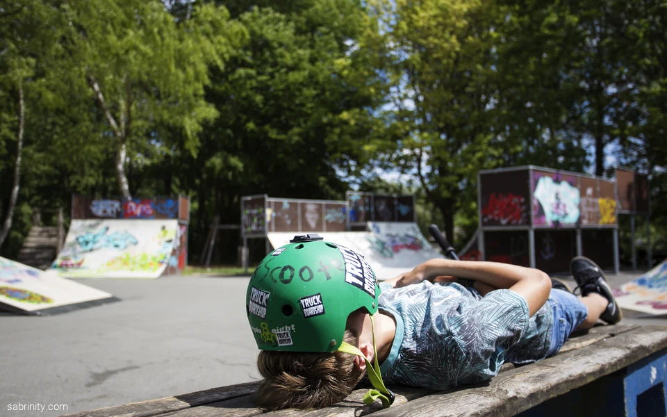 Junge Person mit grünem Helm liegt auf einer Bank in einem Skatepark.